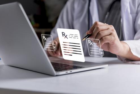 A prescriber holding a pen types on a laptop as multiple digital Rx prescription icons appear projected above the keyboard